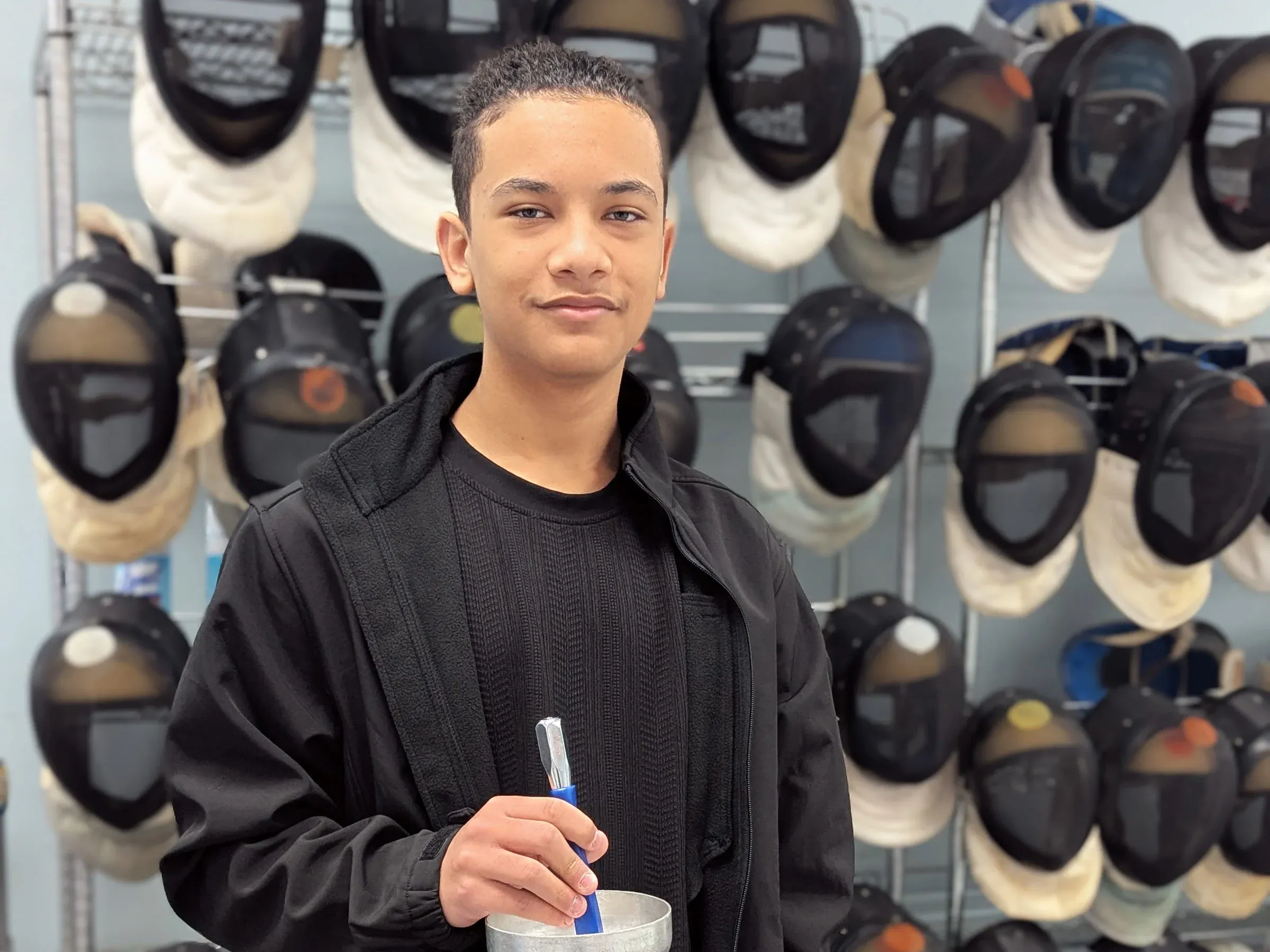 Teen boy smiles at camera holding an epee with French grip. He's wearing a black sweater with black jacket.