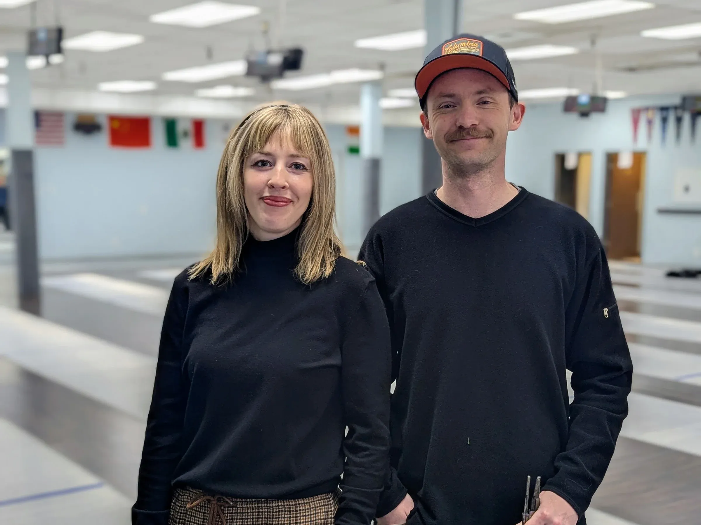 A woman with blond hair and black shirt stands next to a man with a hat in a black shirt. Both are smiling. Fencing club is in the background.